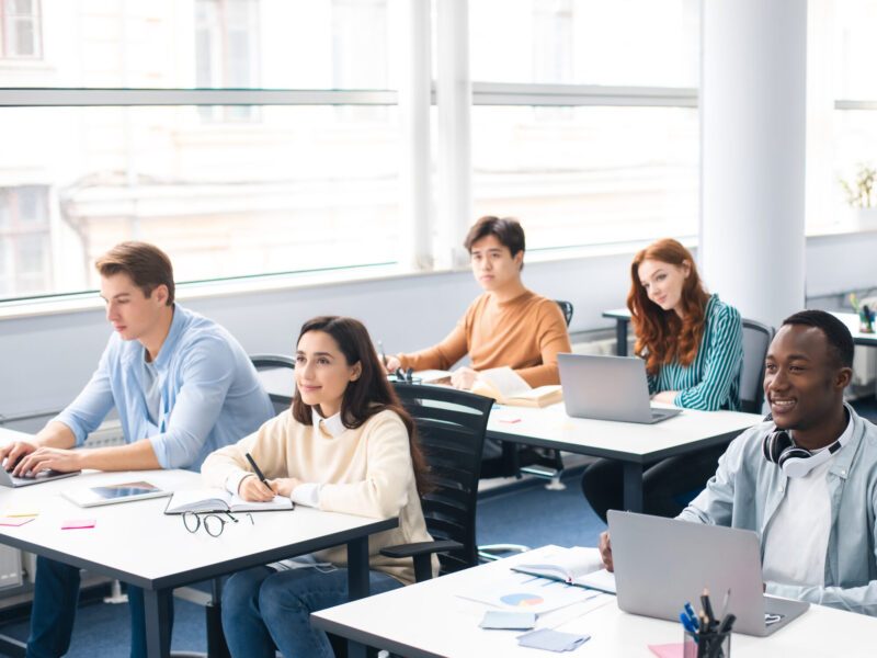 Lecture Concept. Group of interested multicultural mixed race group of students sitting at tables with pc in modern classroom, listening to teacher, taking notes writing in notebooks, back to study